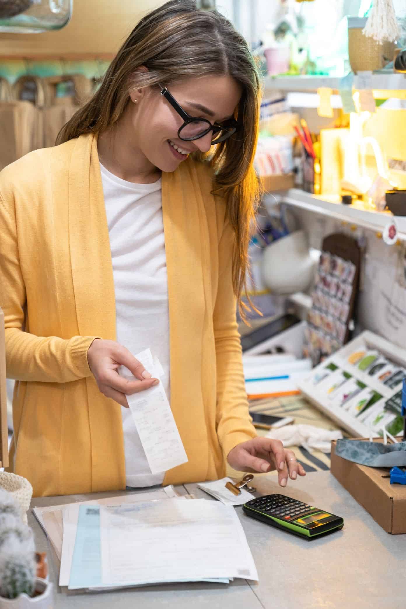 smiling-small-business-owner-in-casual-wearing-yellow-cardigan-standing-in-her-store-using-calculator_t20_G0KvL3
